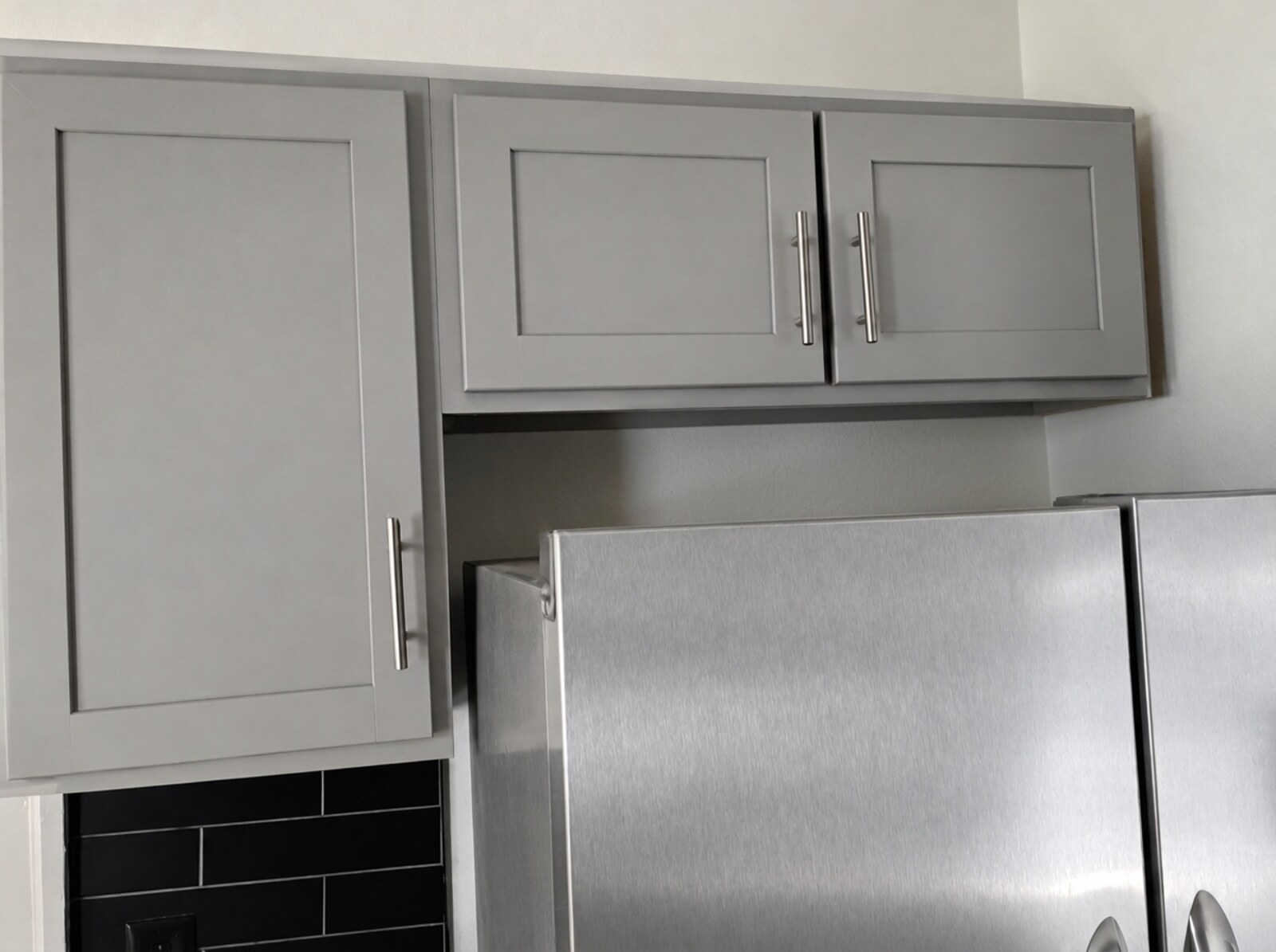 Gray painted upper cabinets above black subway tile with a stainless refrigerator after refinishing
