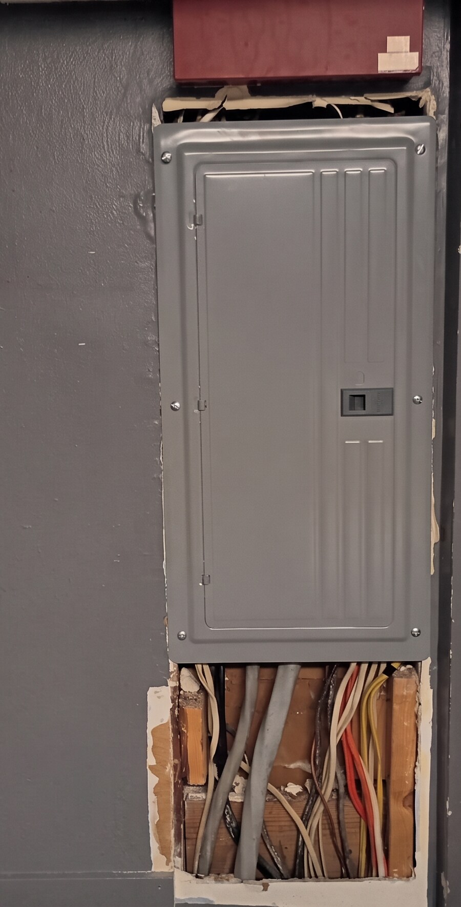 Interior gray wall with the drywall cut away below an electrical breaker panel, exposing wood studs, wiring and conduit, before the repair