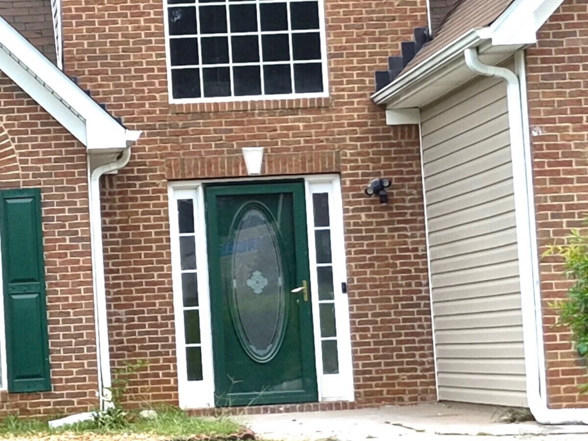 Front entry with a forest green door with an oval glass insert and two white multi-pane sidelights set in a red brick facade, before replacement