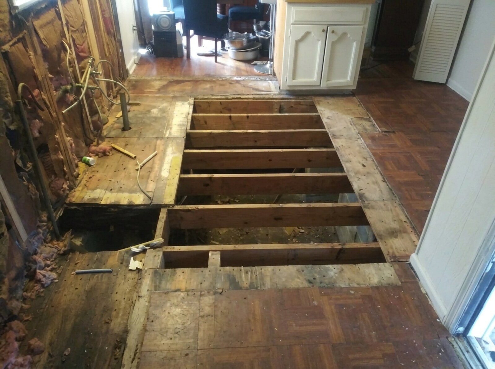 Kitchen mid-demolition with the floor torn out to exposed wood joists and an open hole to the crawl space, wall framing and rough plumbing exposed