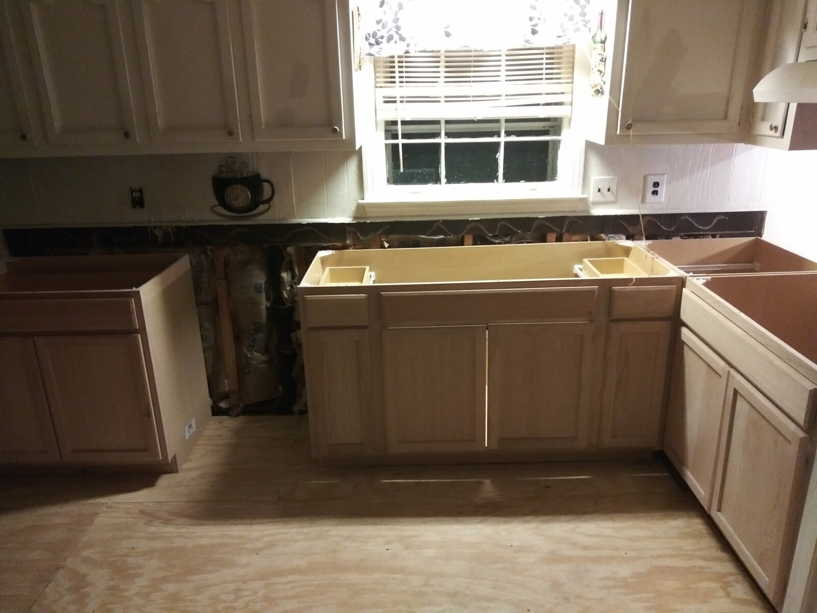 The same kitchen with new plywood subfloor laid and raw unfinished base cabinet boxes installed beneath the existing upper cabinets, wall open for plumbing and electrical